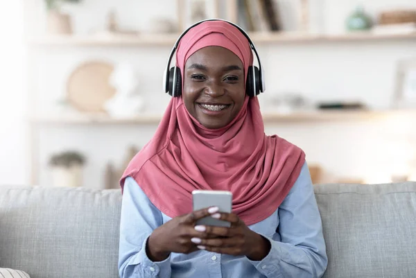 Kesukaan untuk bersantai. Gadis muslim hitam menggunakan smartphone dan headphone nirkabel di rumah — Stok Foto