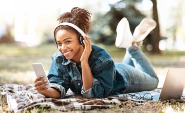 Ekoseli açık havada uzanan kulaklık takan, telefon kullanan Afrikalı kadın.