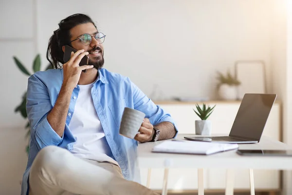 Indio freelancer chico hablando en el teléfono celular y beber café en casa oficina — Foto de Stock