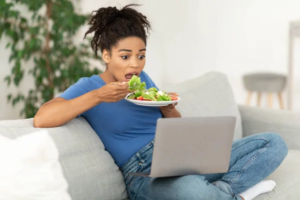 Αφρικανική κοπέλα έχοντας δείπνο βλέποντας ταινία στο φορητό υπολογιστή συνεδρίαση εσωτερικό — Φωτογραφία Αρχείου