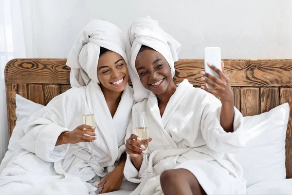 Namoradas deslumbrantes em roupões de banho sentados na cama e tomando selfie — Fotografia de Stock
