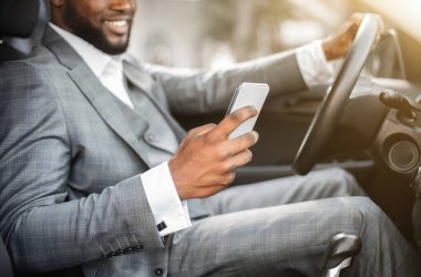 Takım elbiseli siyah adam araba kullanırken akıllı telefon kullanıyor.