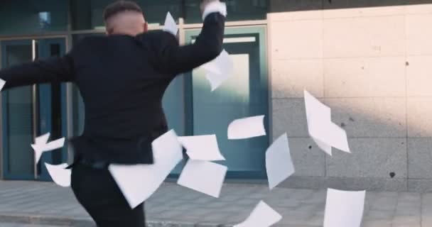 Счастливый бизнесмен в черном костюме танцует и выбрасывает документы на улицу напротив офисного здания — стоковое видео
