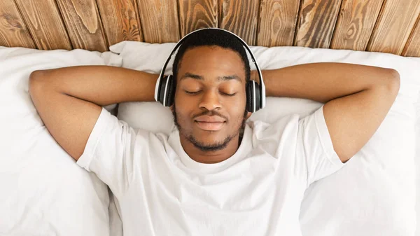 Homem africano ouvindo música usando fones de ouvido deitado na cama dentro de casa — Fotografia de Stock