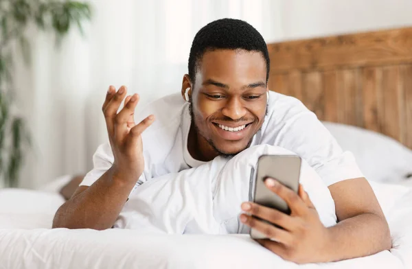 Homem negro feliz fazendo vídeo chamada deitado na cama dentro de casa — Fotografia de Stock