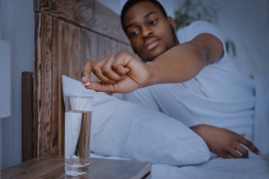 Siyahi adam ilaçlarını alıyor. Evde yatakta yatıyor.