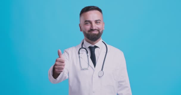医学的承認。陽気な医師ジェスチャー親指アップと笑顔,青のスタジオの背景 — ストック動画
