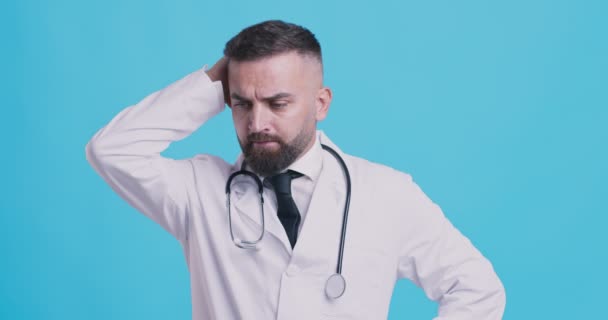 Médico pensativo pensando en el diagnóstico del paciente, arañazos cabeza, fondo estudio azul — Vídeo de stock