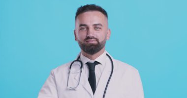 Tıbbi doktor tamam işareti gösteriyor. Mavi stüdyo arka planında gülümsüyor.