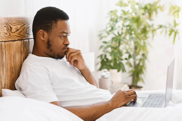 Μαύρος τύπος που χρησιμοποιεί το φορητό υπολογιστή που εργάζονται στο κρεβάτι στο σπίτι — Φωτογραφία Αρχείου