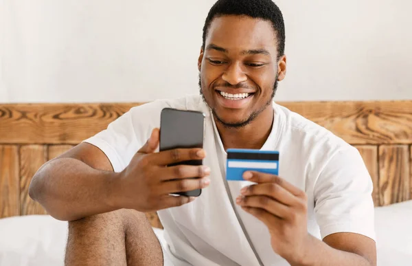 Hombre africano usando Smartphone y tarjeta de crédito sentado en el dormitorio —  Fotos de Stock