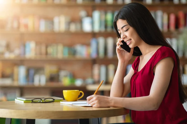 Sourire asiatique femme parler sur cellulaire et prendre des notes dans café — Photo