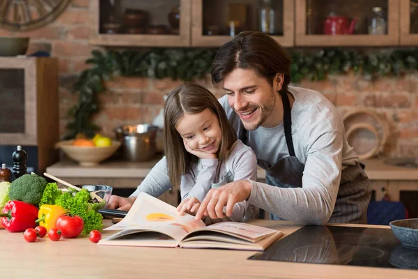 Young Father And Little Daughter Preparing Meal In Kitchen Together, Checking Recipe