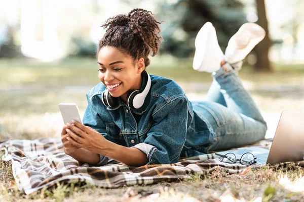 African Millennial Lady za pomocą smartfona leżącego na kratę w parku — Zdjęcie stockowe