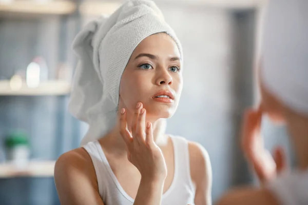 Bella giovane donna controllando la sua pelle per rughe vicino specchio in bagno — Foto Stock