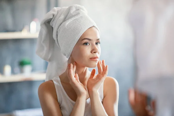 Dame sérieuse en serviette de bain regardant son visage dans le miroir à la salle de bain — Photo