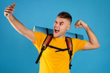 Turist ekipmanlarıyla havalı genç adam mavi stüdyoda akıllı telefondan selfie çekiyor.