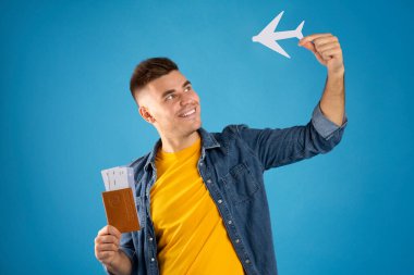 Mavi stüdyo arka planında kağıt uçak, bilet ve uluslararası pasaport olan olumlu milenyum adamı.