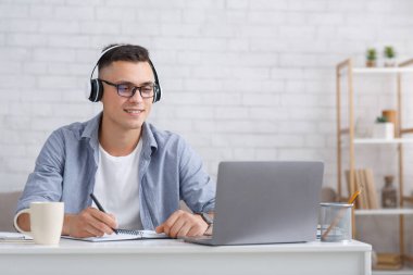 Çevrimiçi webinar ya da covid-19 sırasında ders. Gözlüklü ve kulaklıklı çekici adam dizüstü bilgisayara bakar ve not defterine not alır.