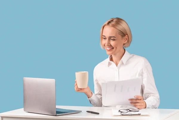 Señora de negocios positiva que trabaja con documentos y beber café frente a la computadora portátil en el escritorio, fondo azul — Foto de Stock