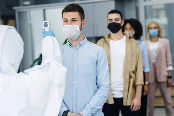 Homme en combinaison de protection mesure la température des travailleurs dans les masques de protection à l'intérieur du bureau, arrêter la propagation de covide-19 — Photo