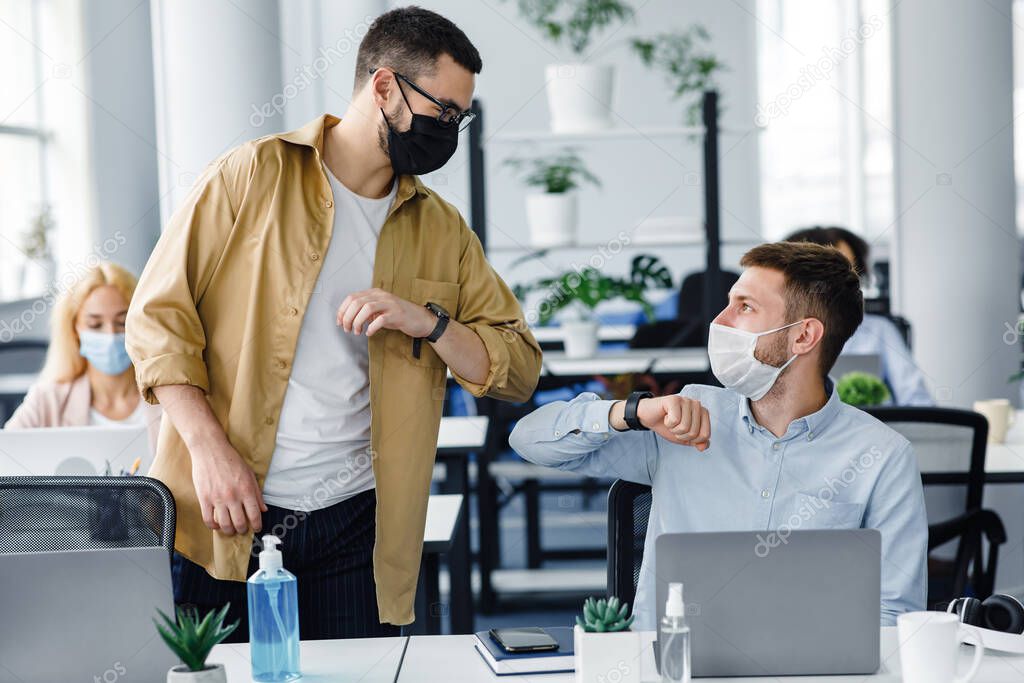 Safe greeting at work during COVID-19 epidemic. Young men in protective masks greet with elbows at workplace with laptop and antiseptic in office