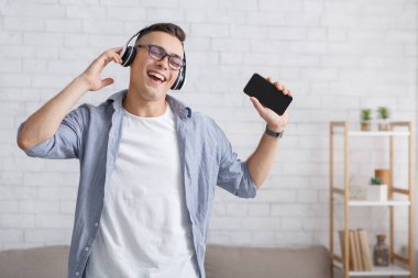 Şarkı söylemek için modern uygulamalar ve koronavirüs salgını sırasında boş zaman. Neşeli adam kulaklıkla şarkı söylüyor ve boş ekranla akıllı telefonu tutuyor.