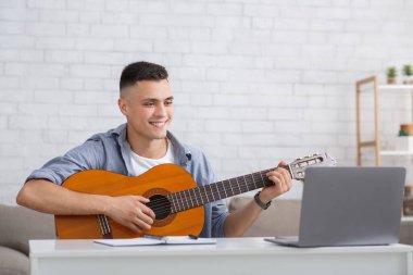 Evde müzik dersi ve COVID-19 salgını sırasında boş zaman. Gitarlı mutlu adam dizüstü bilgisayara bakar ve çalışır.