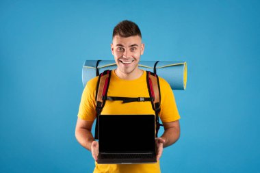 Genç gezgin mavi arka planda boş ekranlı dizüstü bilgisayar gösteriyor. Çevrimiçi turizm ajansı web sitesi için model