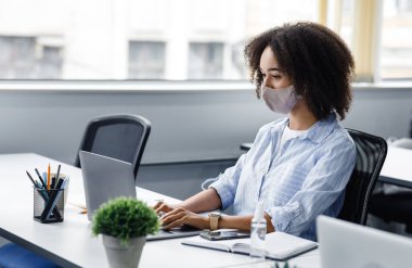 Yeni normal ve ofis işi koruyucu maske ve antiseptikle. Modern iç mekanlarda müşteri siparişleri yazmak için dizüstü bilgisayarda yazan Afrikalı Amerikalı kadın.