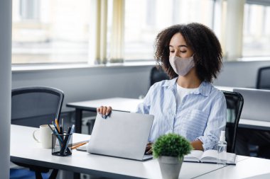 Günün sonunda eve git. Koruyucu maskeli Afrikalı Amerikalı kadın laptopu kapatıyor, modern ofis içinde antiseptikle masada oturuyor.