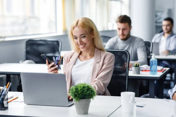 El gerente trabaja con el cliente en línea. Trabajadora sonriente sosteniendo teléfono inteligente y escribiendo en el portátil en el lugar de trabajo en el interior de la oficina moderna — Foto de Stock