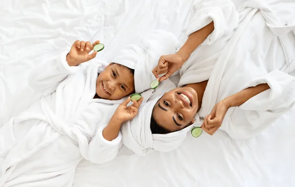 Spa casero. Feliz mamá africana y su hija en batas de baño con rodajas de pepino — Foto de Stock