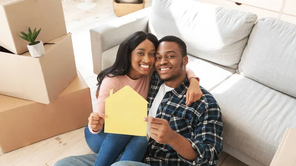 Negro millennial chico con su novia abrazando y sosteniendo casa de papel en el suelo de su nuevo apartamento en día de mudanza — Foto de Stock