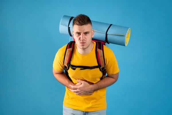 Upset male tourist with camping equipment suffering from stomach pain on blue studio background
