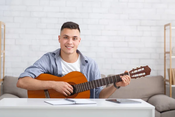 Online music lessons. Cheerful guy with guitar sits on couch in living room interior and records video or makes record for modern blog — Stock Photo, Image