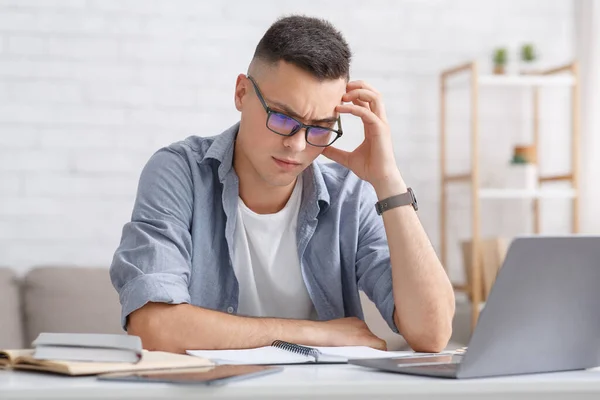 Schwierige Aufgabe und Fernunterricht zu Hause. Brainstorming eines jungen Mannes, der mit Laptop am Tisch sitzt und im Wohnzimmer auf das Notizbuch schaut — Stockfoto