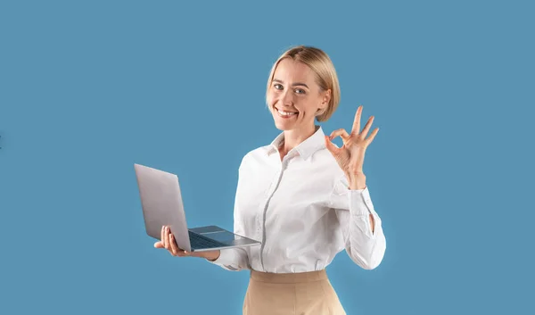穿着正式服装的快乐的金发女商人在蓝色工作室背景的笔记本电脑上工作时表现得很好 — 图库照片