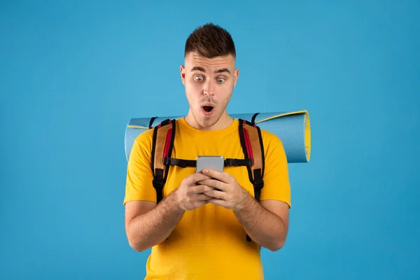 Σοκαρισμένος αρσενικό τουρίστας με το smartphone κράτηση περιοδεία σε απευθείας σύνδεση, έκπληκτος πάνω από μεγάλη έκπτωση διακοπών πάνω από μπλε φόντο — Φωτογραφία Αρχείου