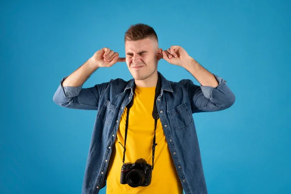 Ερεθισμένος άνδρας ταξιδιώτης με φωτογραφική μηχανή που καλύπτει τα αυτιά του με δάχτυλα σε μπλε φόντο στούντιο — Φωτογραφία Αρχείου