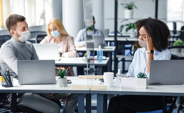 Sichere Kommunikation am Arbeitsplatz und soziale Distanz während der covid-19. Kollegen reden durch Schutzglas, Afroamerikanerin mit Maske leidet unter Kopfschmerzen — Stockfoto