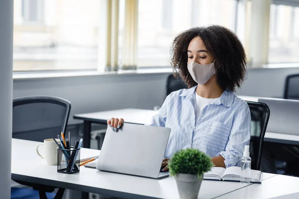 End of day and go home. African american woman in protective mask closes laptop, sitting at table with antiseptic in modern office interior — Stock Photo, Image