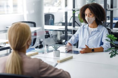 Koruyucu maskeli genç işçiler COVID-19 enfeksiyonunu önlemek için bölme yoluyla birbirleriyle konuşuyorlar.