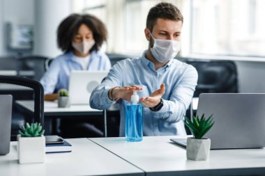 Koruyucu maskeli genç adam dizüstü bilgisayarda çalışıyor ve ellerini antiseptikle dezenfekte ediyor.