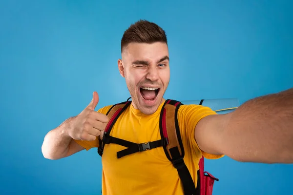 Joven hipster fresco con equipo turístico tomando selfie, guiñando el ojo y mostrando gesto pulgar hacia arriba sobre fondo azul — Foto de Stock