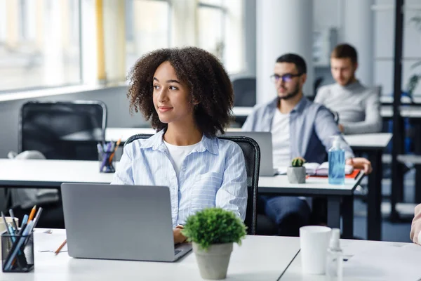 Creación de procesos de negocio e ideas de gestión. Mujer afroamericana mirando a la ventana en el lugar de trabajo, trabajando en el ordenador portátil en el interior de la oficina moderna con colegas — Foto de Stock