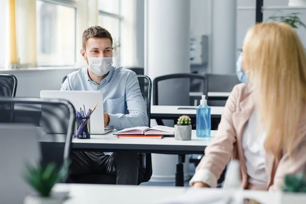 Social distance of workers in office during quarantine. Guy and woman in protective masks are talking, sitting at tables with computers and antiseptics — Stock Photo, Image