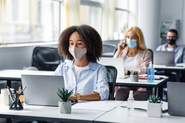 Social distancing at coronavirus outbreak situation. Pretty african american lady in protective mask typing on laptop, working for company support center — Stock Photo, Image