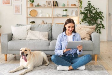 Evde elinde tablet ve köpekle genç bir kadın var.