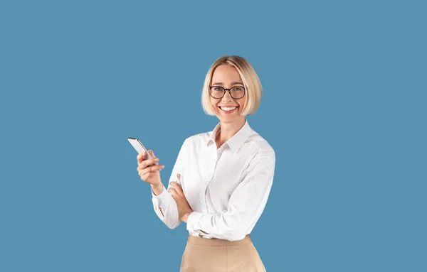 Pewna siebie bizneswoman ze smartfonem uśmiechnięta i patrząca w kamerę na tle niebieskiego studia — Zdjęcie stockowe